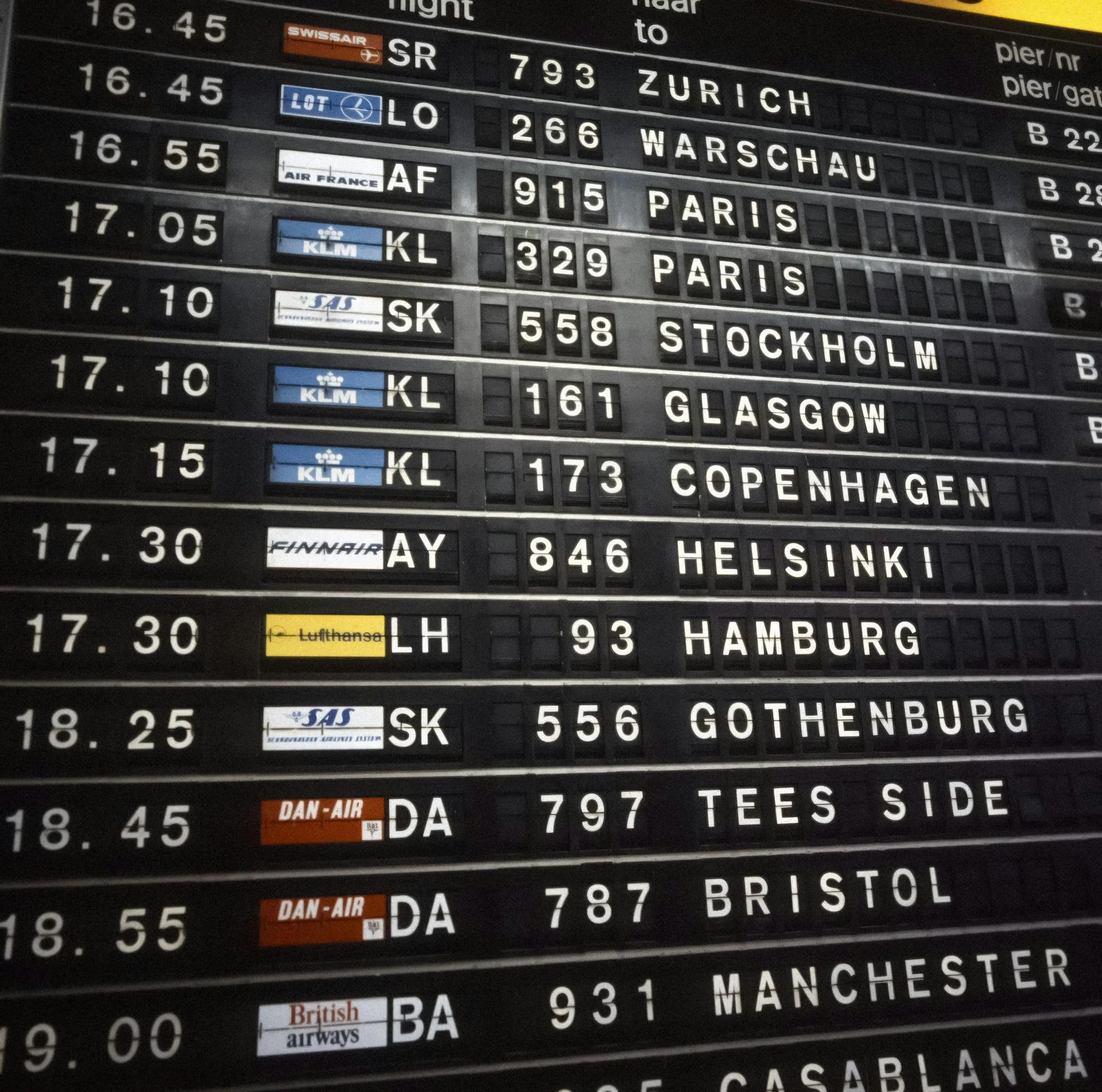 A split-flap departures board at Schiphol airport in the 1980s, rows of white letters on black listing flights to Stockholm, Glasgow, Copenhagen, Helsinki and Hamburg